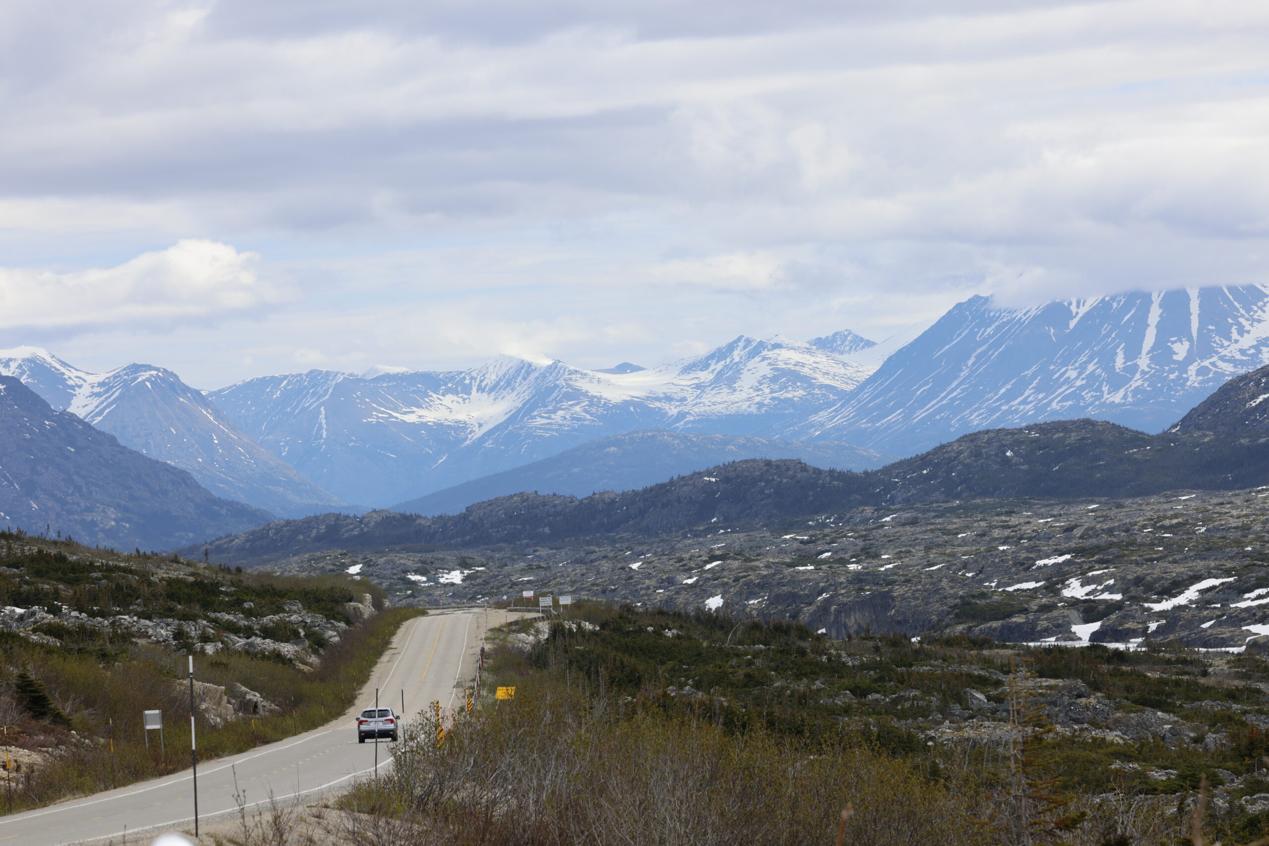 winding road leads through a mountainous landscape with snow-capped peaks and cloudy skies,