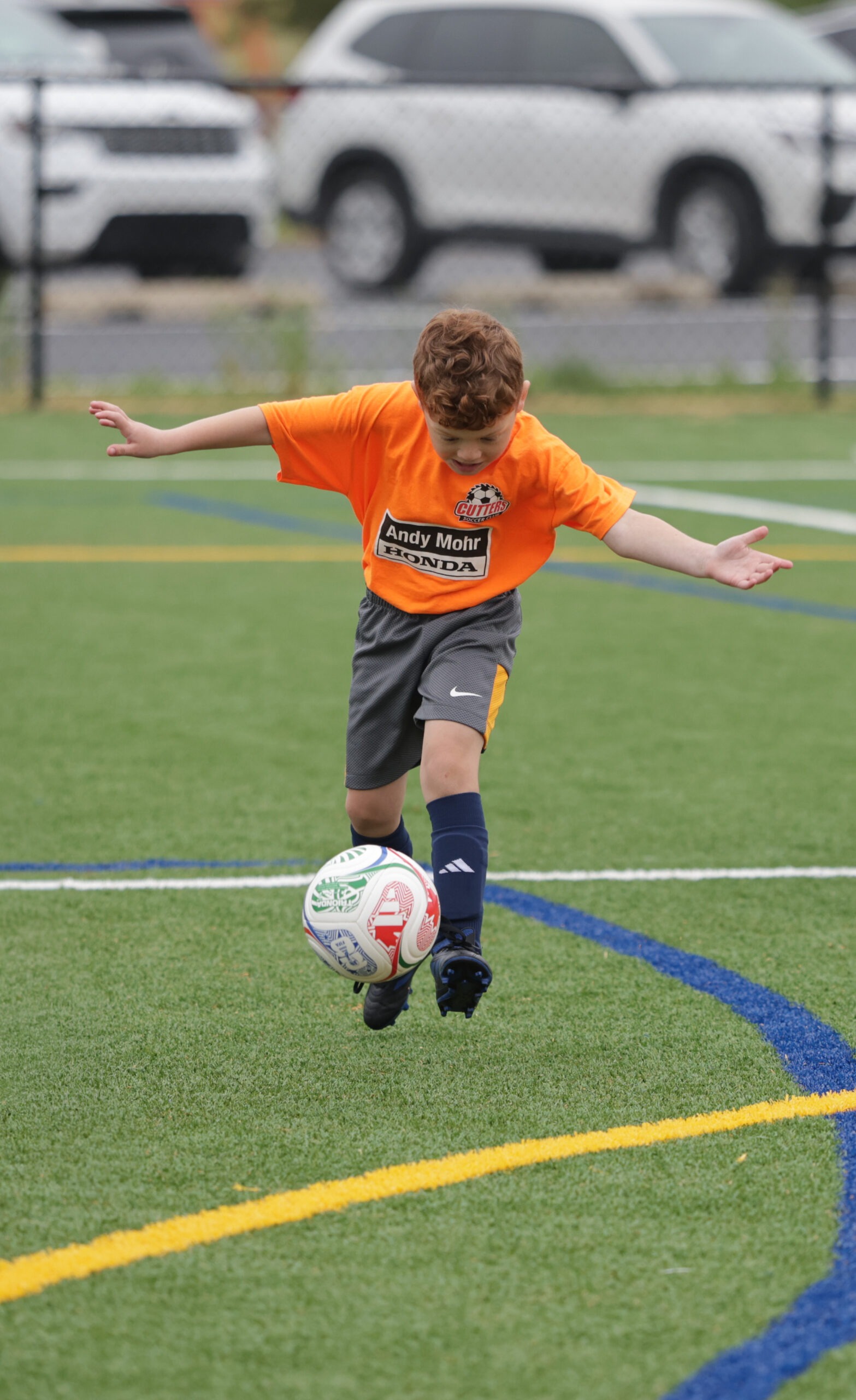 A young boy in an orange soccer jersey skillfully dribbles a multicolored soccer ball on a vibrant green field